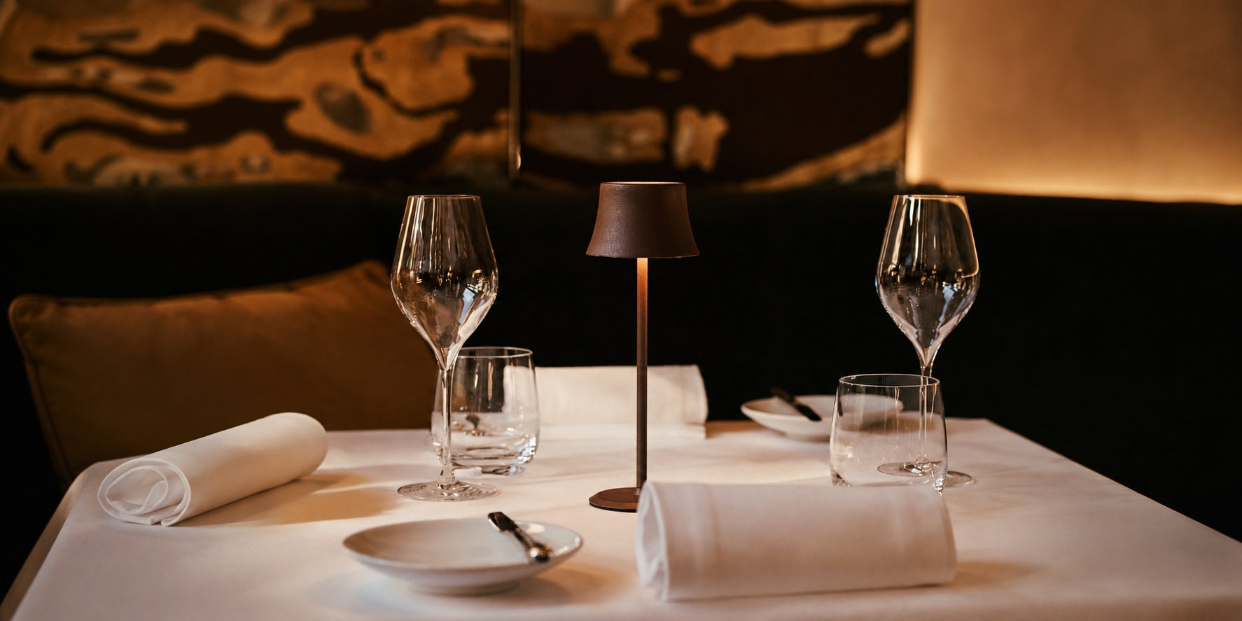 Tavellio bronze wireless table lamp glowing on a white-clothed fine dining restaurant table with wine glasses.