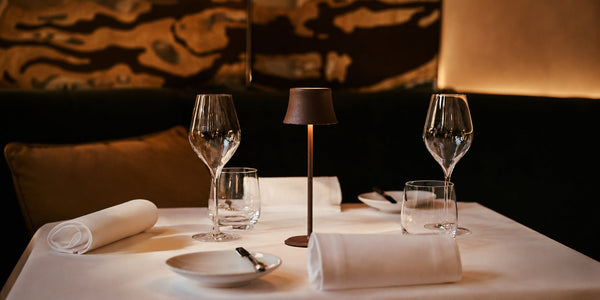 Tavellio bronze wireless table lamp glowing on a white-clothed fine dining restaurant table with wine glasses.