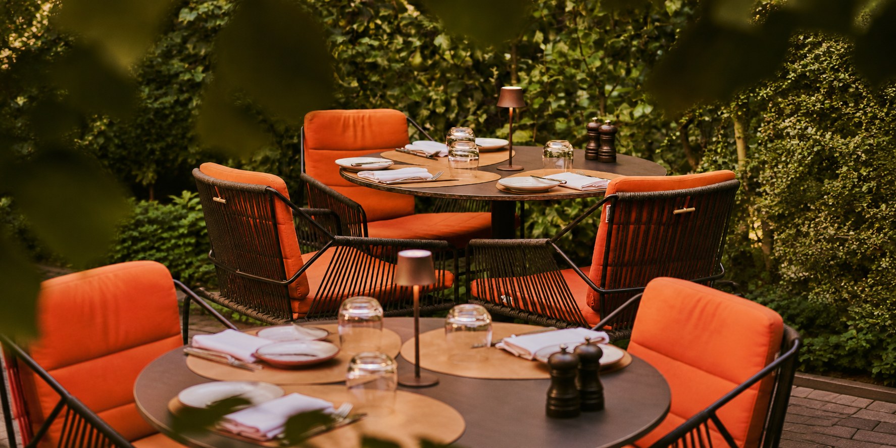 Tavellio small wireless table lamps softly lighting restaurant terrace tables surrounded by lush green hedges.