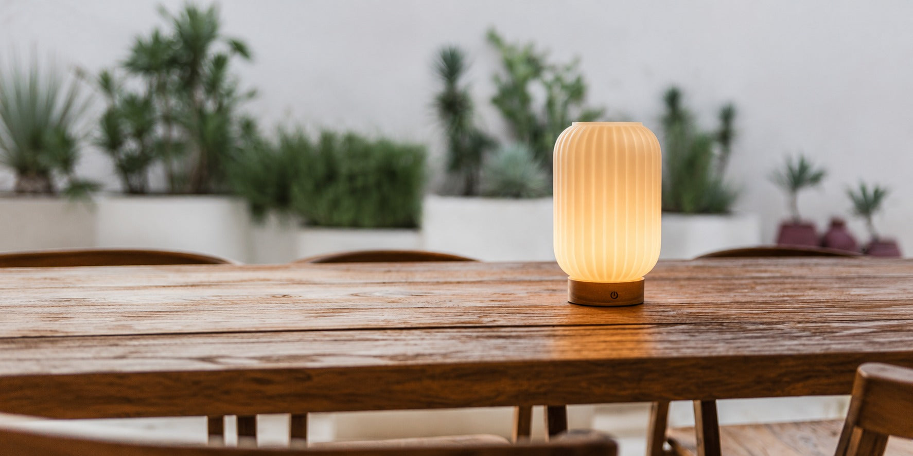 Tavellio fluted portable table lamp glowing warmly on a wooden outdoor dining table surrounded by potted plants.
