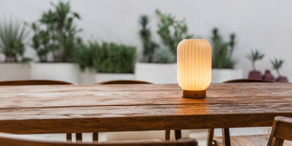 Tavellio fluted portable table lamp glowing warmly on a wooden outdoor dining table surrounded by potted plants.