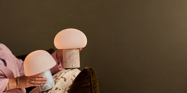 Two glowing rechargeable table lamps with marble bases held in hand against a dark olive wall on an autumn evening