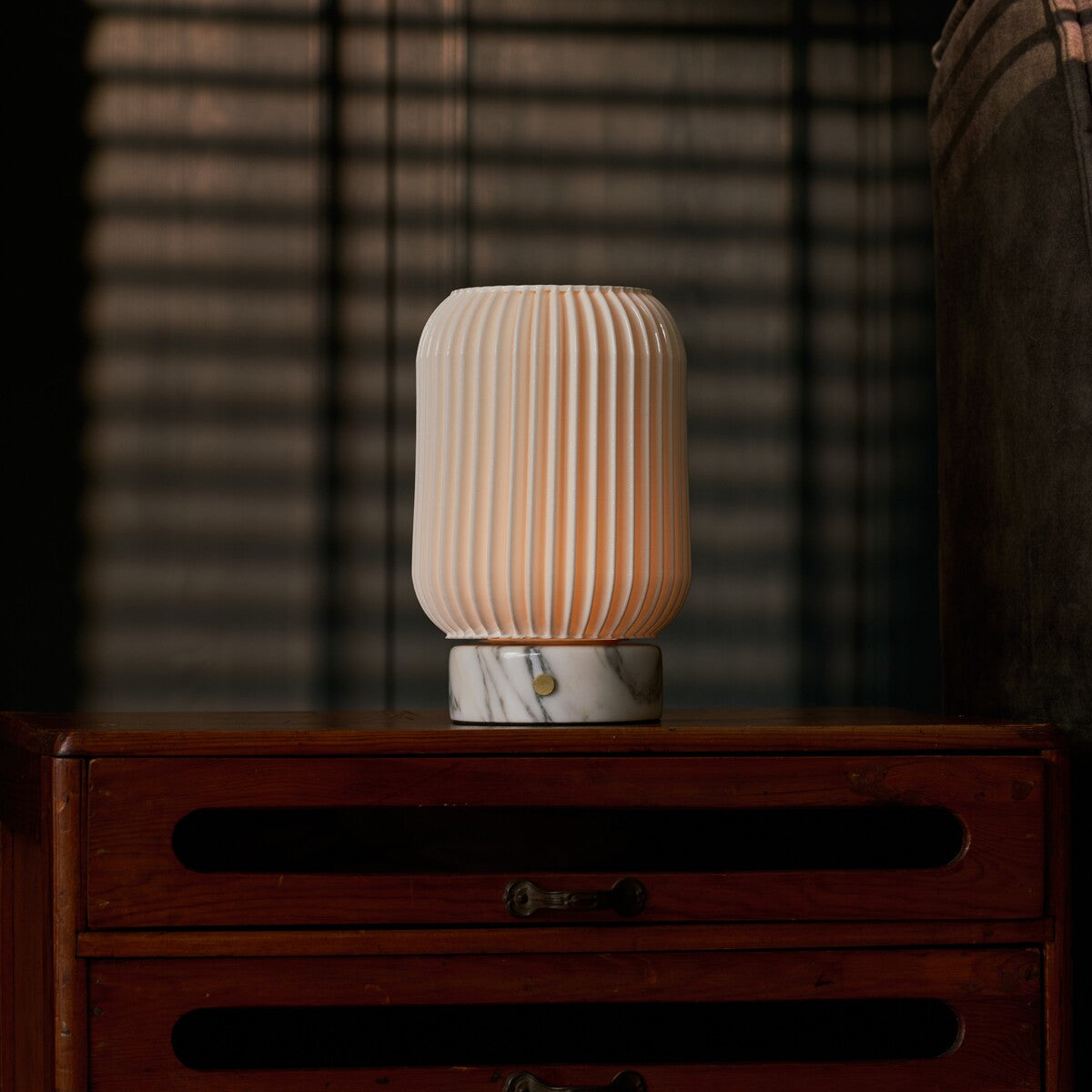 Rechargeable table lamp Alba lit on a wooden dresser with blinds-cast shadows in a bedroom