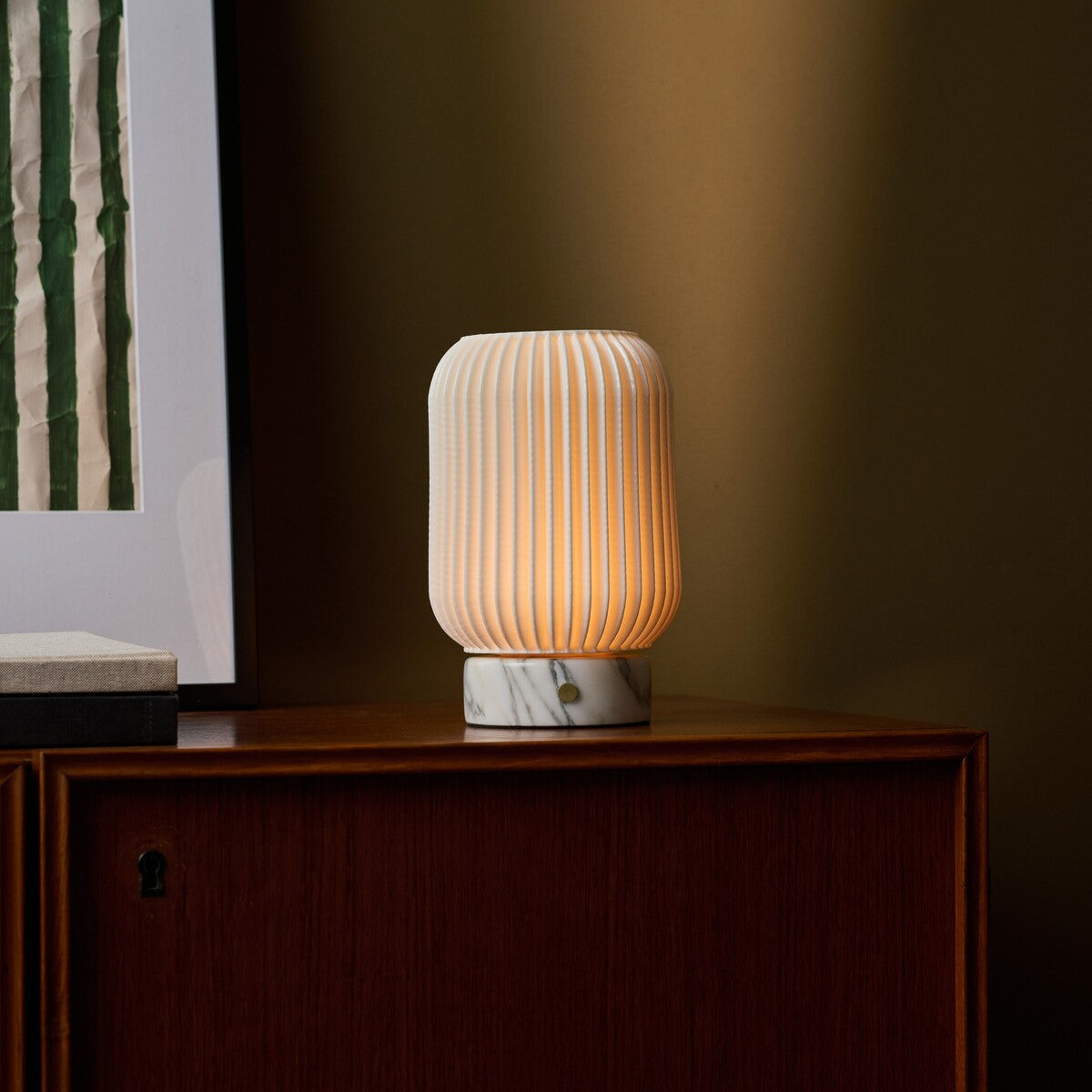 Tavellio rechargeable table lamp Alba glowing on a wooden sideboard beside striped framed art