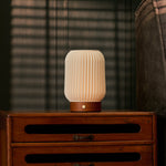 Cordless table lamp Alba in chestnut brown wood on a rustic nightstand beside a striped armchair