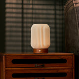 Cordless table lamp Alba in chestnut brown wood on a rustic nightstand beside a striped armchair