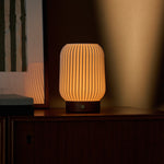 Cordless table lamp Alba in chestnut brown wood glowing warmly beside a framed botanical print
