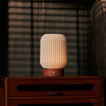 Cordless table lamp Alba in red travertine and porcelain, glowing on a bedroom nightstand beside striped blinds