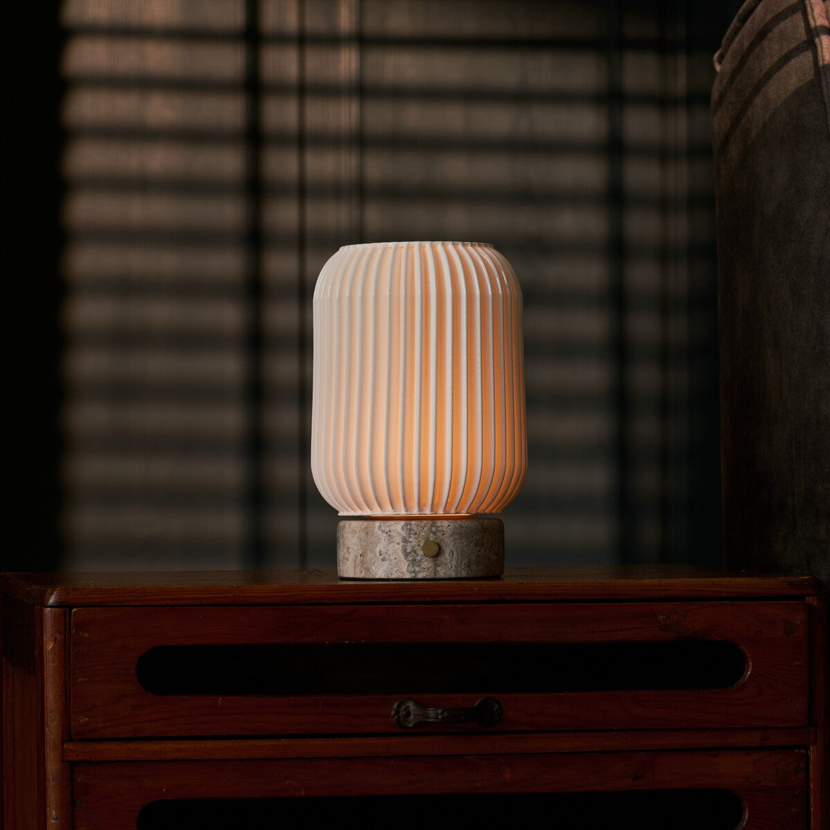 Alba rechargeable table lamp in silver travertine and porcelain, glowing on a bedroom nightstand beside striped blinds