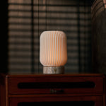 Alba rechargeable table lamp in silver travertine and porcelain, glowing on a bedroom nightstand beside striped blinds