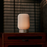 Alba rechargeable table lamp in silver travertine and porcelain, glowing on a bedroom nightstand beside striped blinds