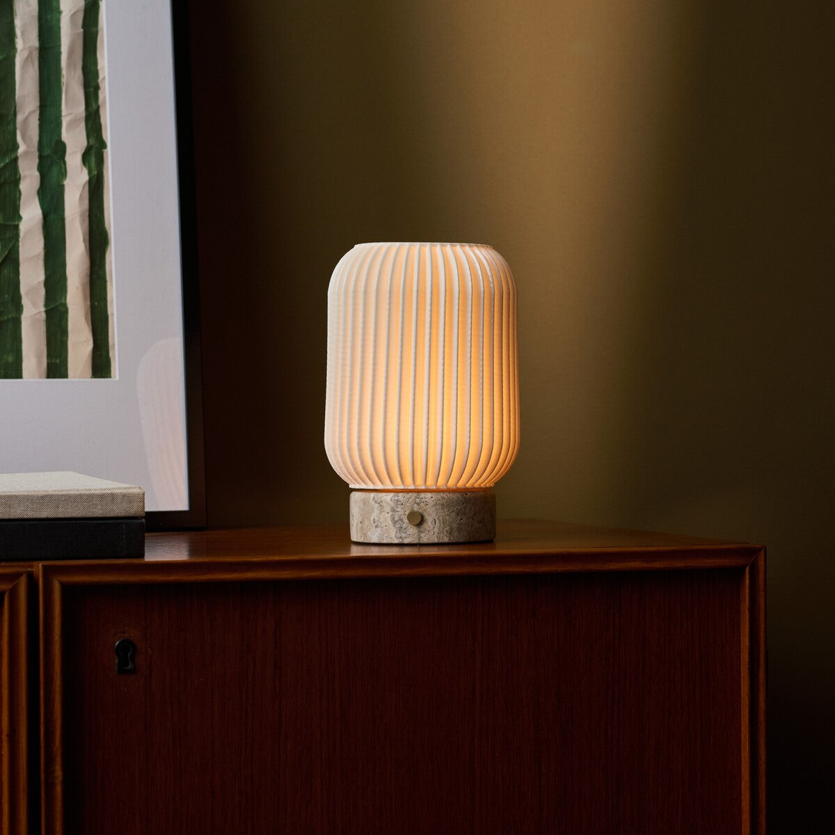 Tavellio Alba cordless table lamp in silver travertine and porcelain, warm glow beside framed botanical artwork