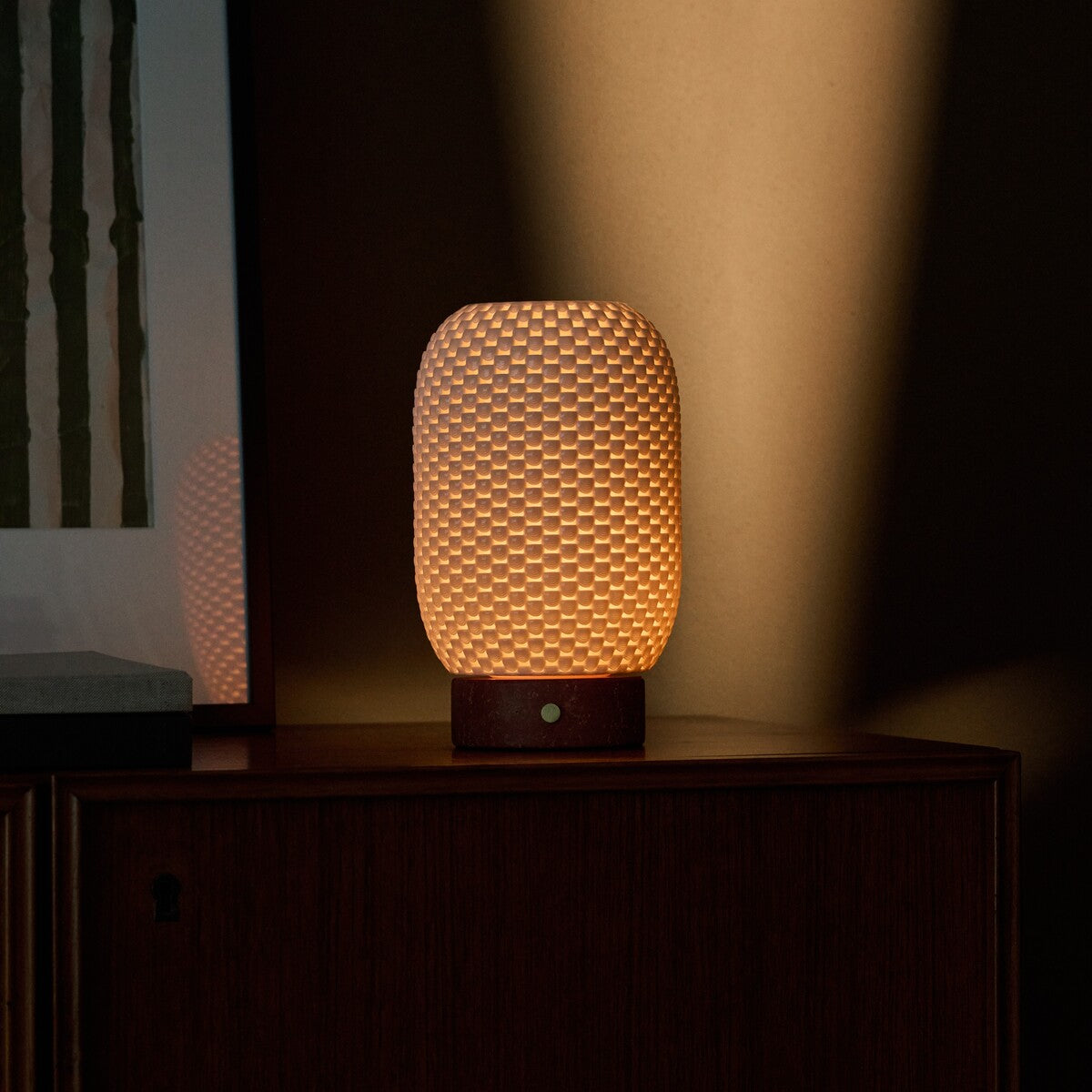 Cordless table lamp Aurora in Red Travertine and porcelain casting warm light reflected in a nearby picture frame