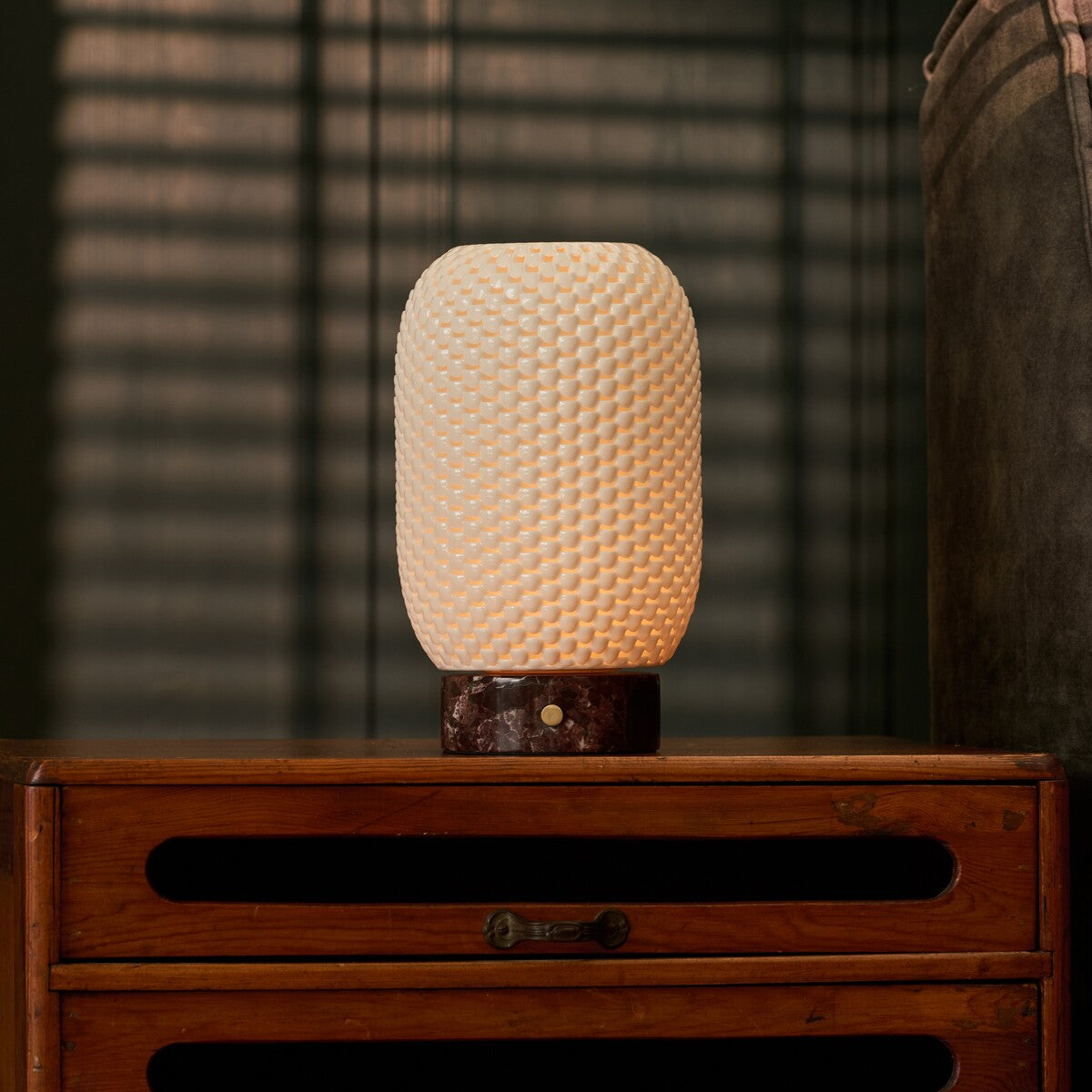 Aurora rechargeable table lamp glowing on a wood dresser, blinds casting striped shadows