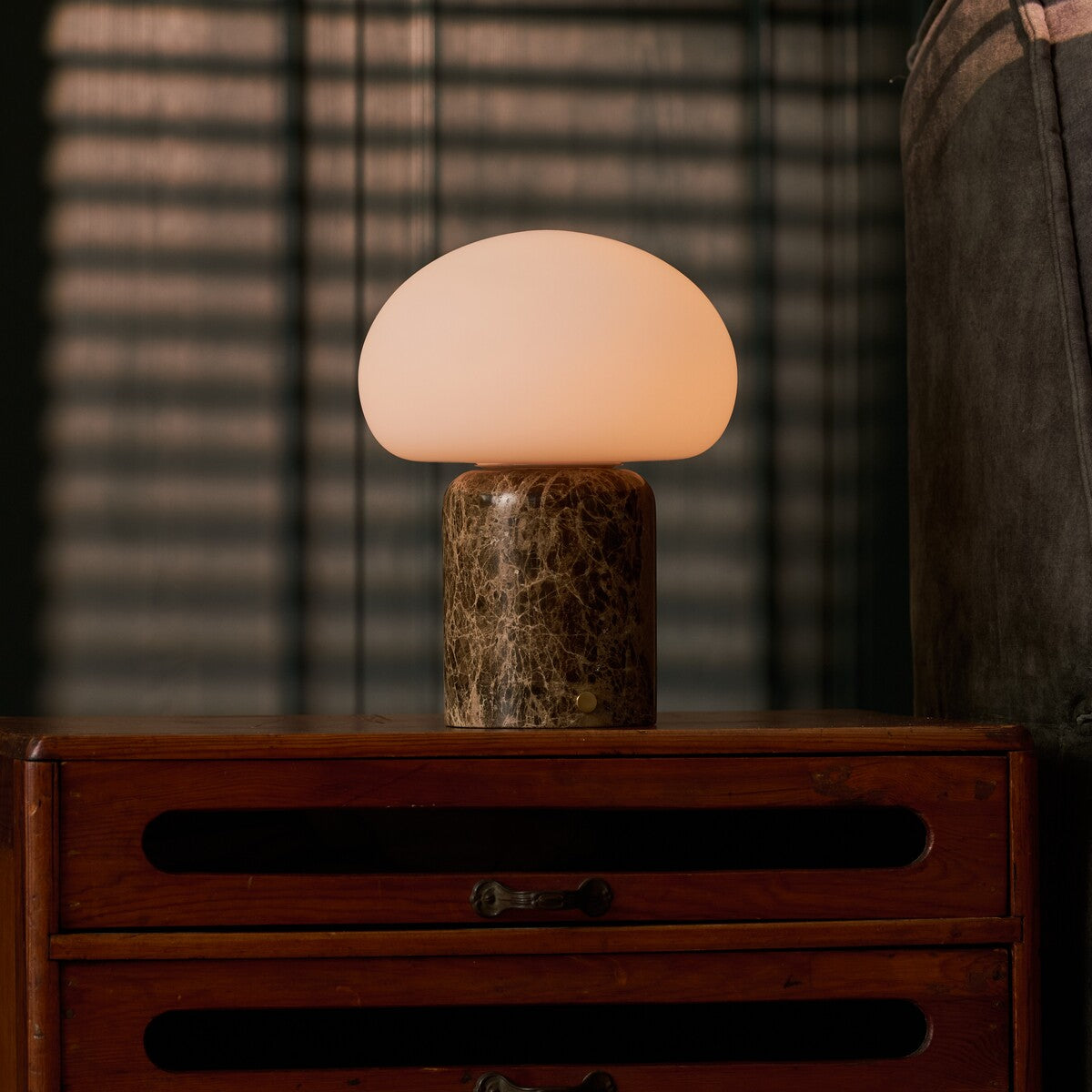Coddé Large marbre Emperador foncé lampe de table sans fil Tavellio – commode en bois foncé devant des stores vénitiens – coussin rayé visible