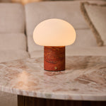 Rechargeable table lamp with red travertine base glowing on a round stone table beside a cream sofa