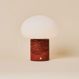 Rechargeable table lamp with red travertine base and opal glass dome shade on a plain backdrop