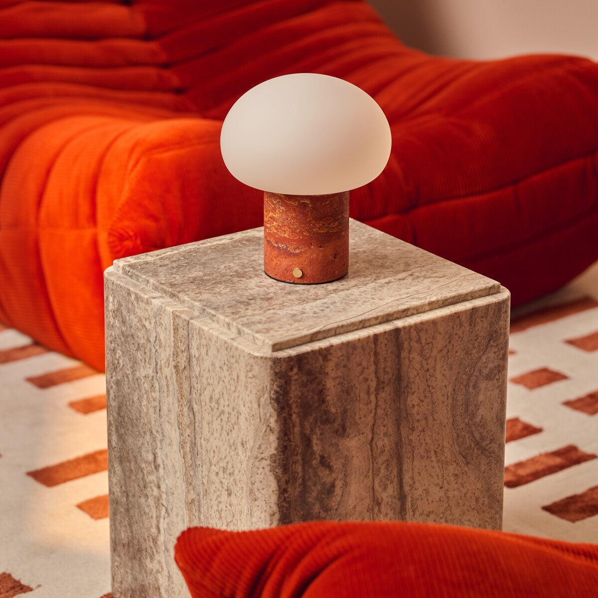 Rechargeable table lamp with red travertine base on a travertine plinth with an orange corduroy cushion in view