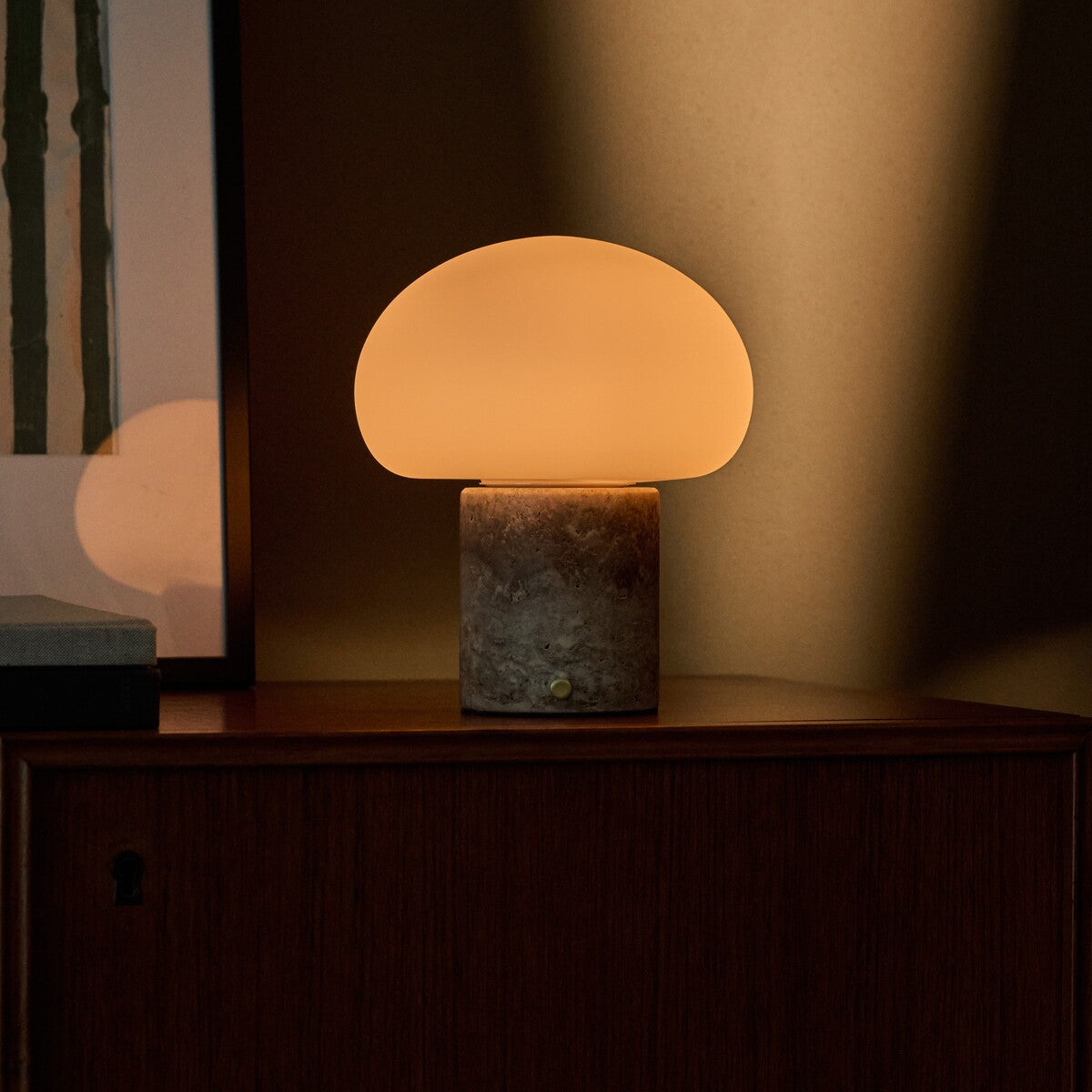 Cordless table lamp glowing atop a walnut sideboard, its travertine base catching warm evening light