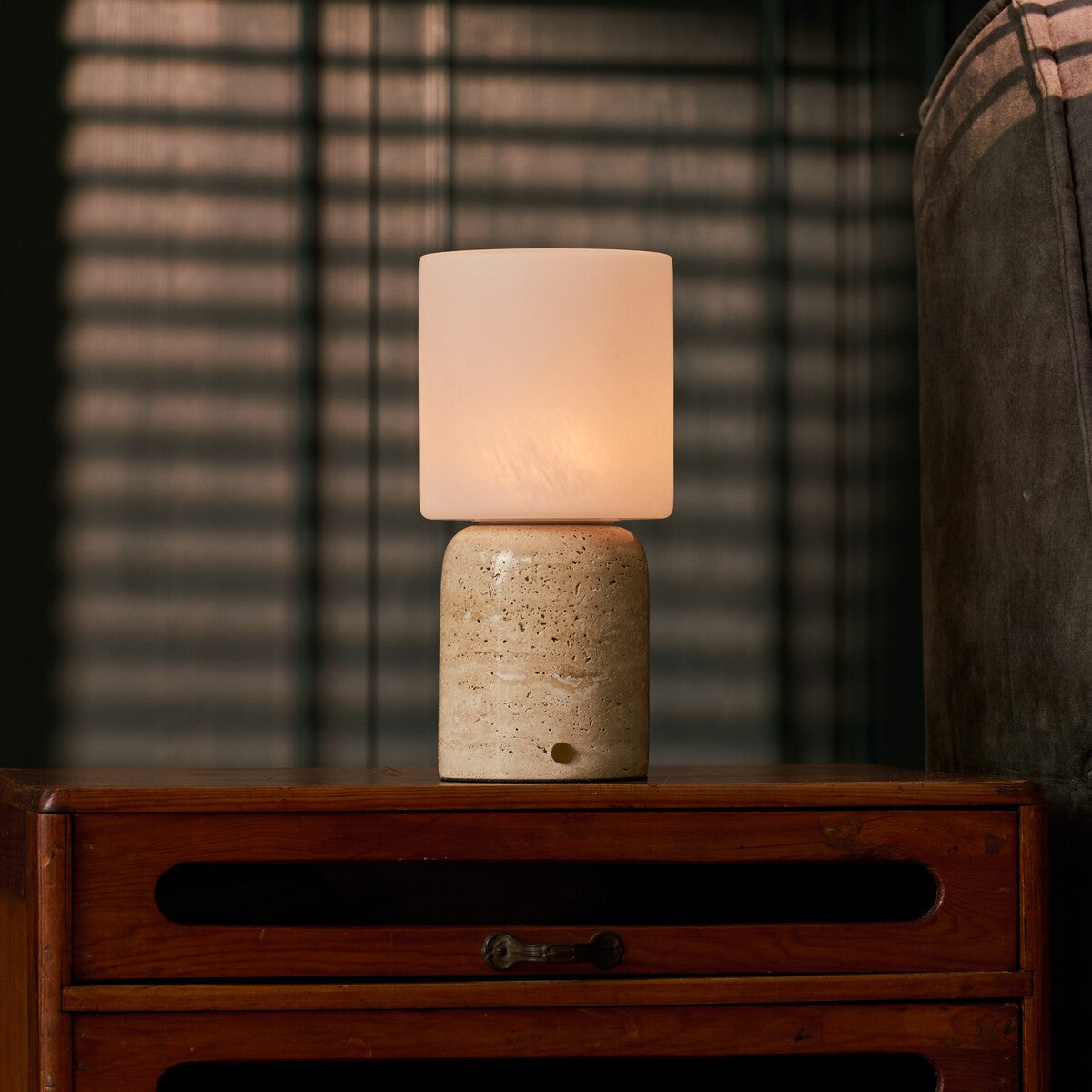 Cuyp Large cordless table lamp with beige travertine base glowing on a nightstand beside striped blinds