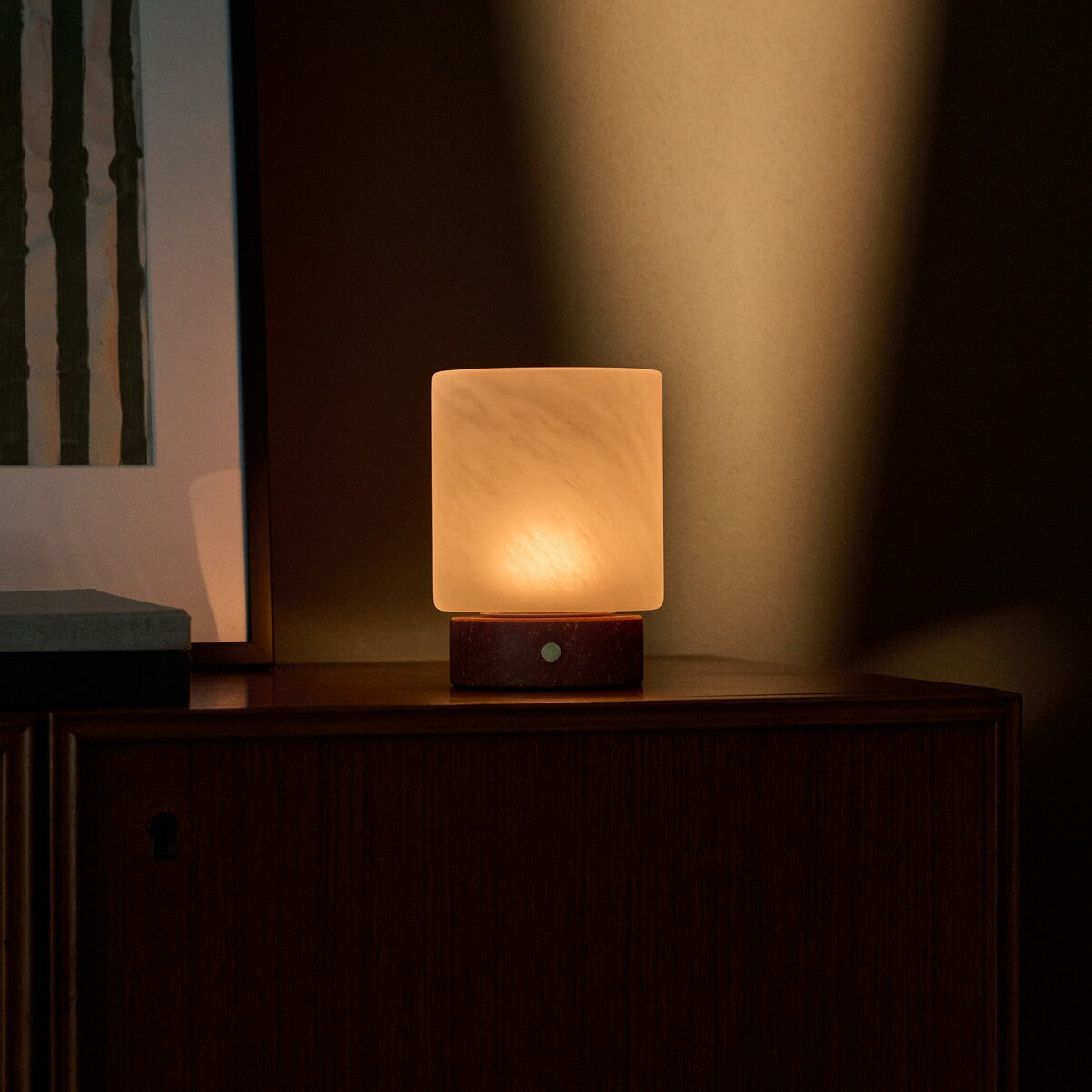 Lampe de table Cuyp Small travertin rouge sans fil Tavellio - allumée sur un meuble en bois près d'un cadre à rayures