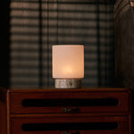 Cordless table lamp with travertine base lit softly on a dark dresser beneath striped blind shadows