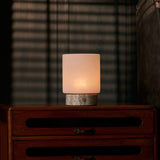 Cordless table lamp with travertine base lit softly on a dark dresser beneath striped blind shadows