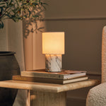 Dou cordless table lamp in Arabescato marble glowing beside stacked books and an olive branch