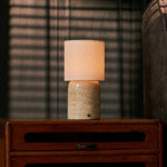 Cordless table lamp Dou Large Classic Travertine on a bedside table with striped blinds and armchair