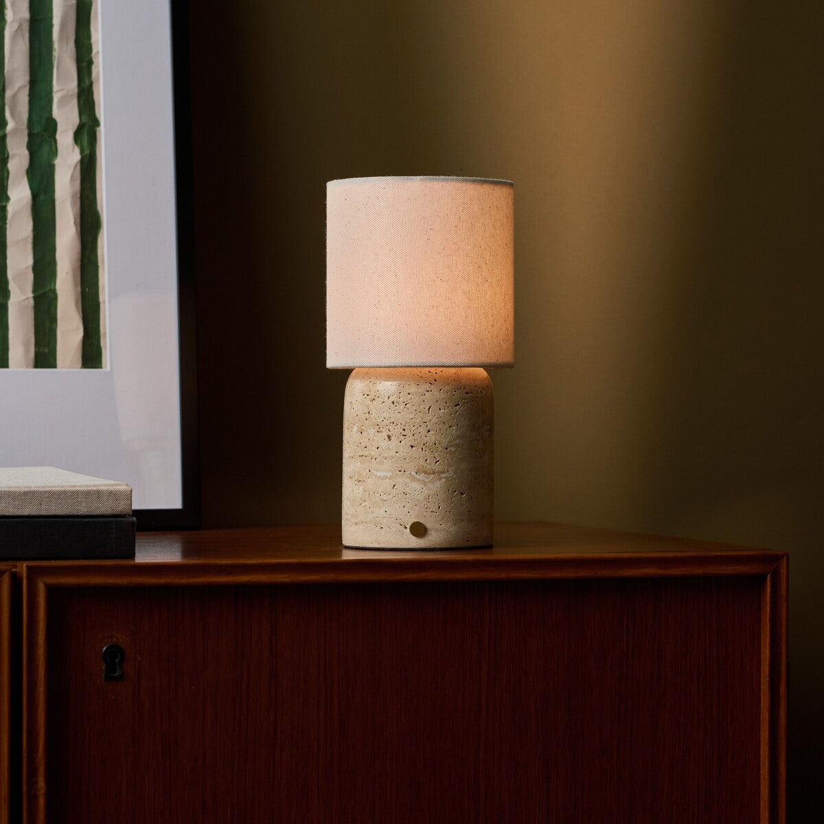 Rechargeable table lamp Dou Large Classic Travertine glowing beside a framed print on a wood cabinet