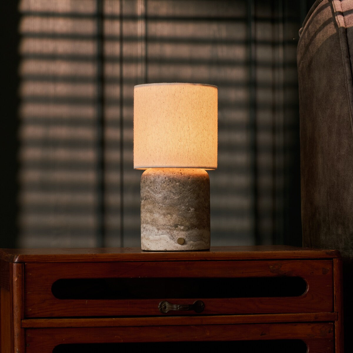 Cordless table lamp Dou Large Silver Travertine glowing on a nightstand beside striped blinds