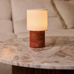 Dou cordless table lamp in red travertine glowing on a marble side table beside a beige sofa