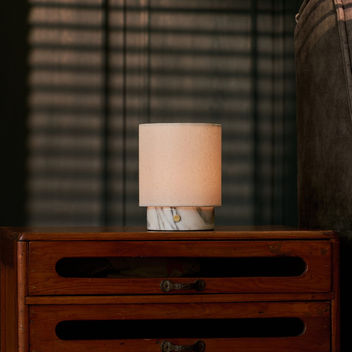 Cordless Dou Small Arabescato Marble Table Lamp glowing on a wooden dresser in front of striped blinds