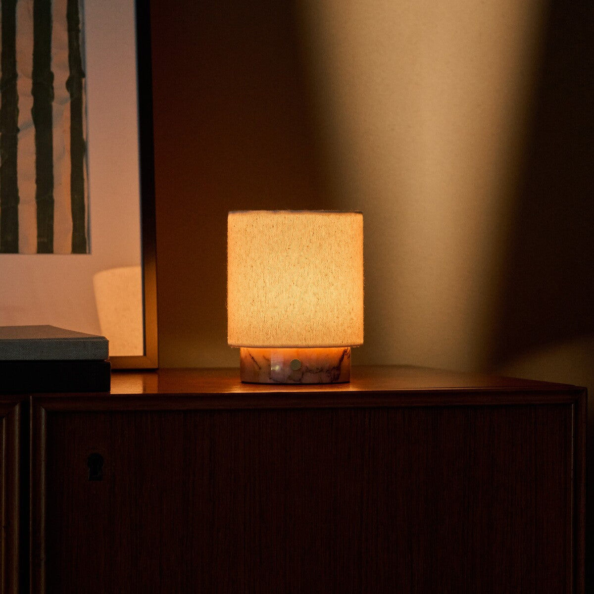 Tafellamp oplaadbaar Dou Small brandt warm op een donkere houten console naast gestreepte wandkunst