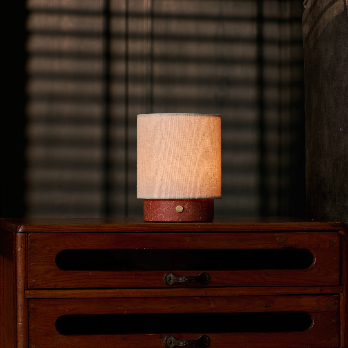 Dou Small rechargeable table lamp in red travertine on a vintage cane-front dresser with blind-shadow light