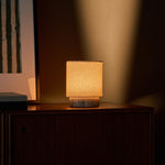 Dou Small Silver Travertine Rechargeable Table Lamp lit on a vintage wooden dresser in front of striped blinds