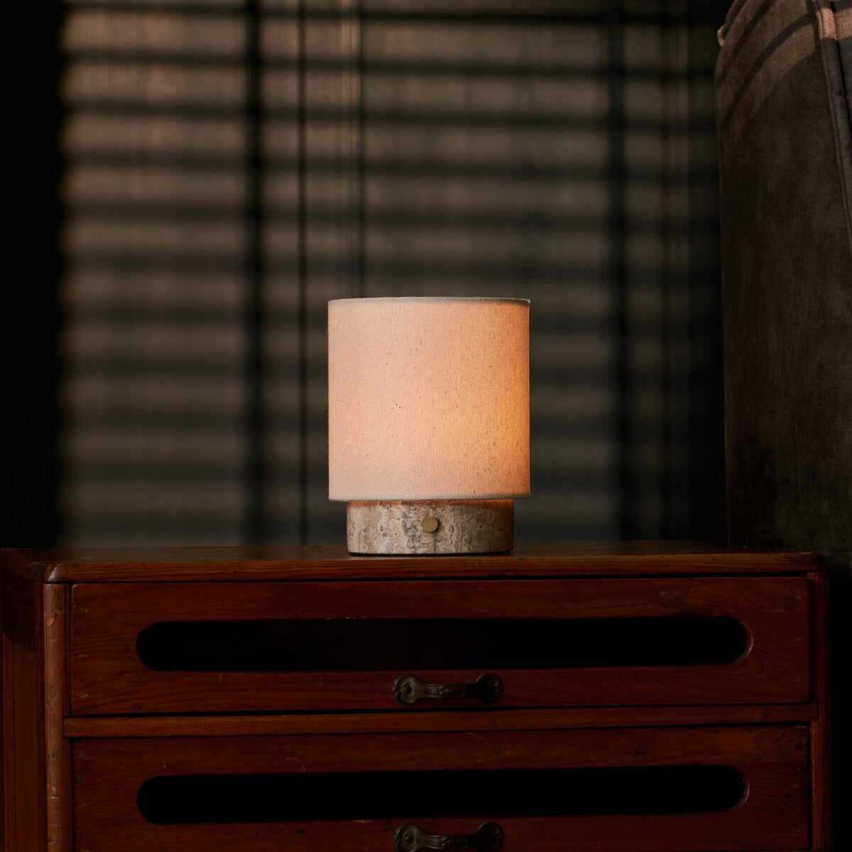 Cordless Dou Small Silver Travertine Table Lamp glowing on a dresser beside a grey upholstered armchair