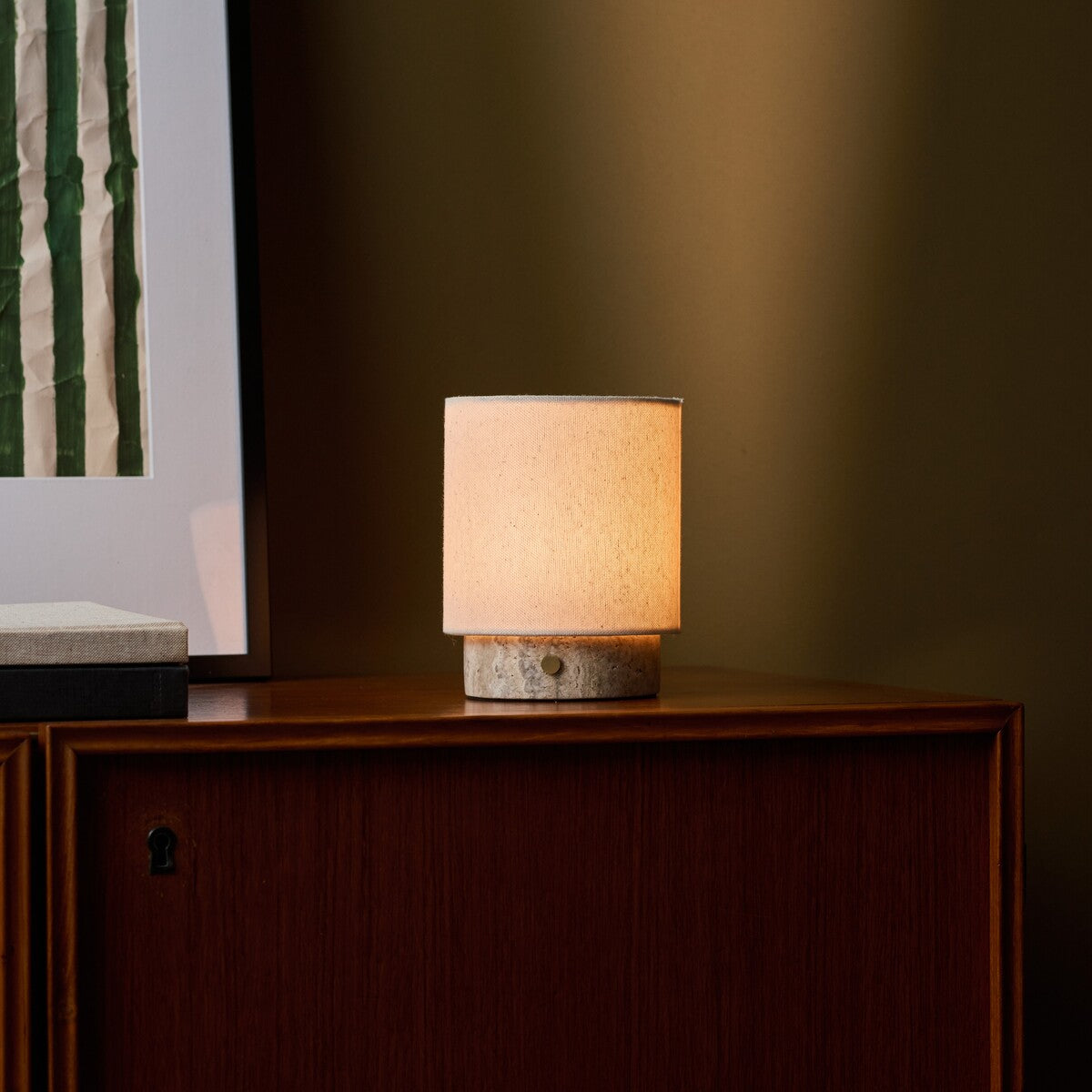 Tavellio Dou Small Silver Travertine Rechargeable Table Lamp lit beside framed art on a walnut cabinet