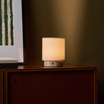 Tavellio Dou Small Silver Travertine Rechargeable Table Lamp lit beside framed art on a walnut cabinet