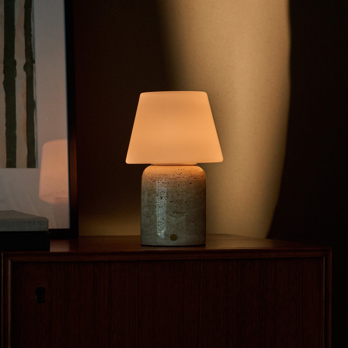 Oplaadbare tafellamp Hooch Large in beige travertin verspreidt warm licht bij schemering op een houten kast