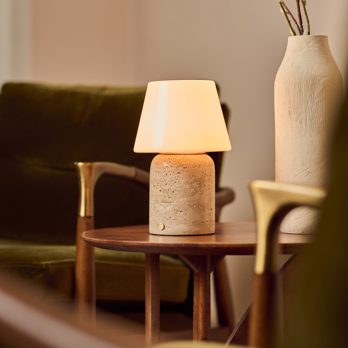 Tafellamp oplaadbaar Hooch Large in beige travertin gloeit op een houten bijzettafel naast een groene bank