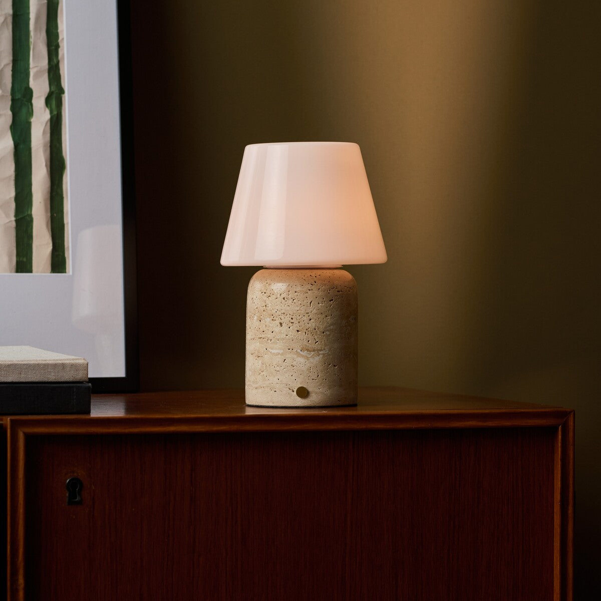 Oplaadbare tafellamp Hooch Large in beige travertin verlicht een donker houten dressoir tegen een olijfgroene muur