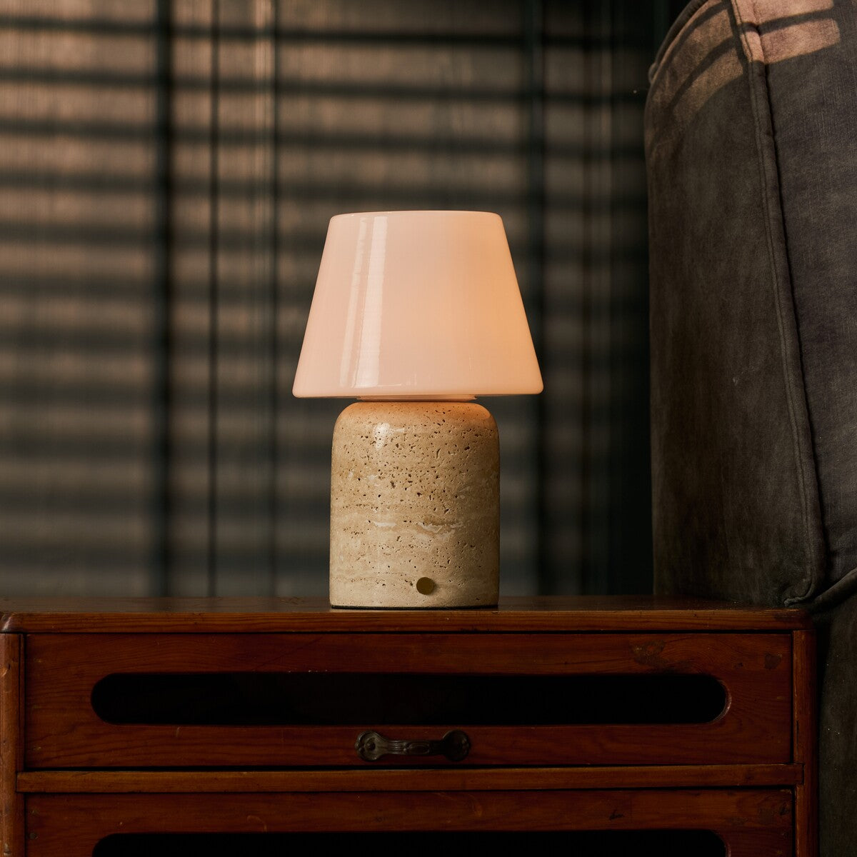 Tafellamp oplaadbaar Hooch Large in beige travertin gloeit op een houten dressoir bij gestreepte jaloezieën