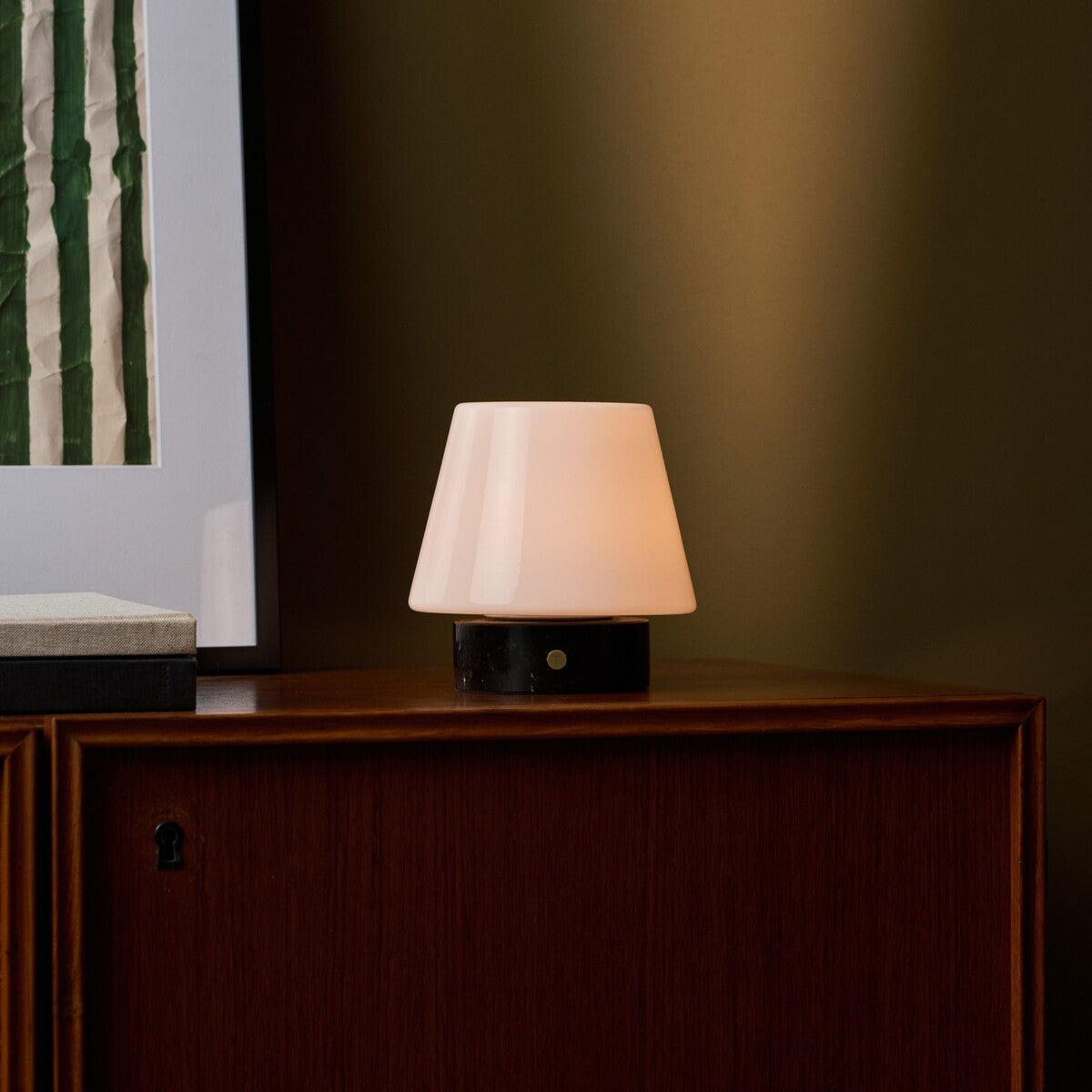 Oplaadbare tafellamp Hooch Small Nero Marquina Marble brandend op een houten kast naast gestreepte wandkunst