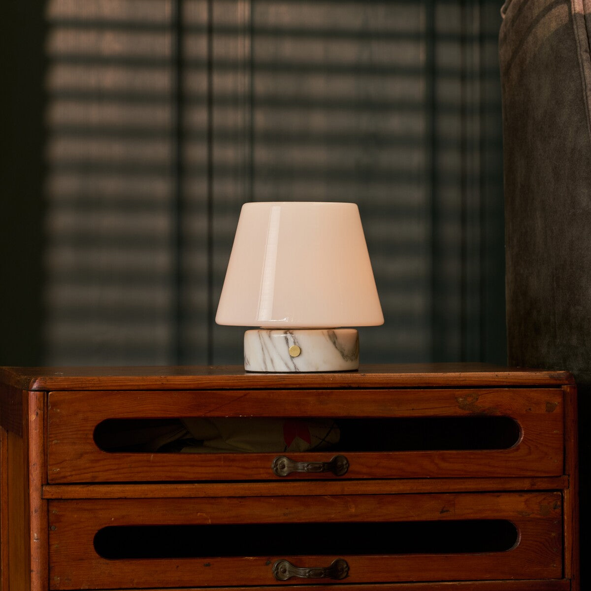Tafellamp oplaadbaar Hooch Small brandend op een vintage houten dressoir bij jaloezieen en een fauteuil