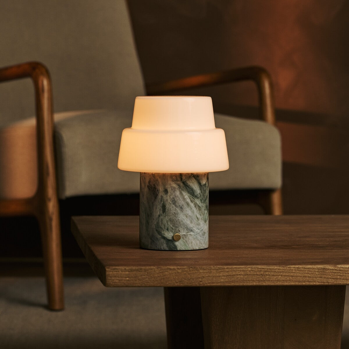 Cordless Pissarro table lamp in ice green marble glowing warmly on a wooden side table between armchairs.