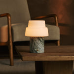 Cordless Pissarro table lamp in ice green marble glowing warmly on a wooden side table between armchairs.