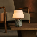 Cordless Pissarro table lamp in ice green marble glowing warmly on a wooden side table between armchairs.