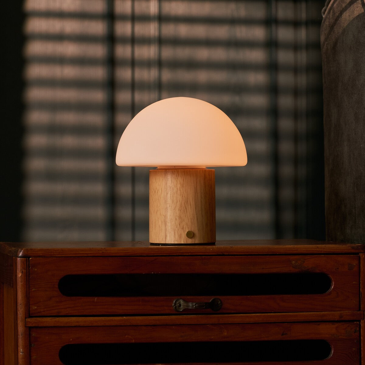 Cordless table lamp Ronald with light brown wood base and frosted glass dome shade on a wooden dresser in front of a sunlit blind
