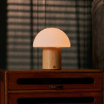 Cordless table lamp Ronald with light brown wood base and frosted glass dome shade on a wooden dresser in front of a sunlit blind